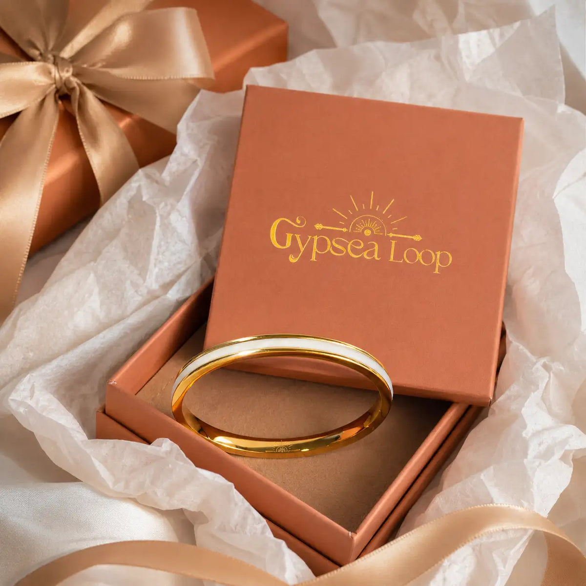 A polished gold bangle bracelet displayed inside an open terracotta-colored jewelry box, with the lid resting behind it. The box is branded 'Gypsea Loop' in gold lettering.